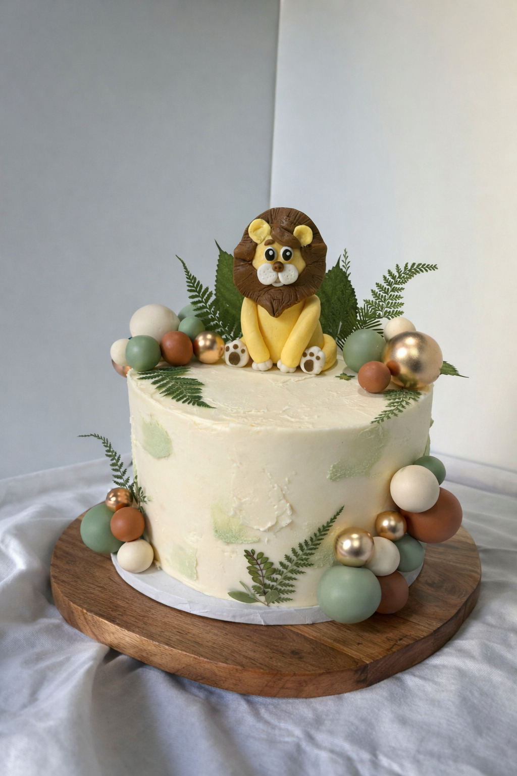 Cream-colored safari cake with a lion topper, fern details, and decorative spheres on a wooden stand.