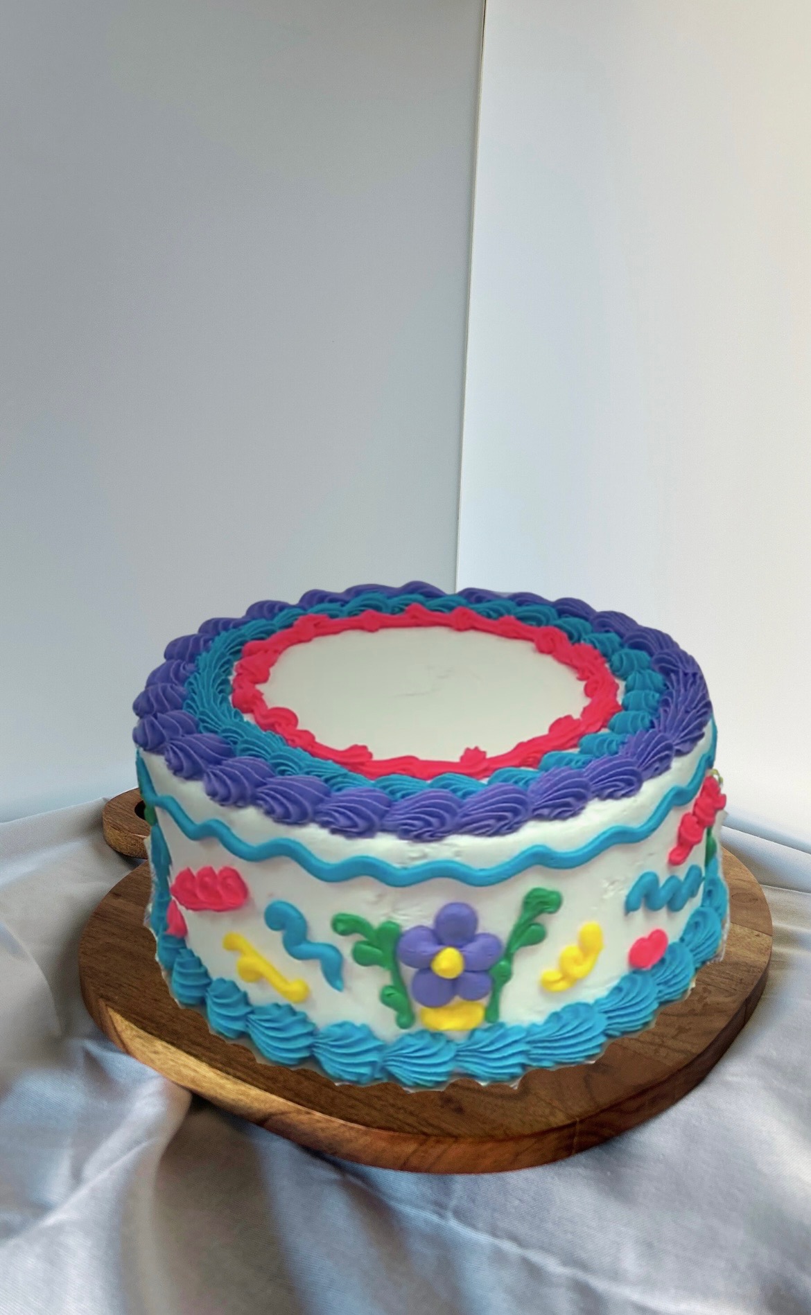 Round vintage-style cake with purple, blue, and pink piping and floral details on a wooden stand.