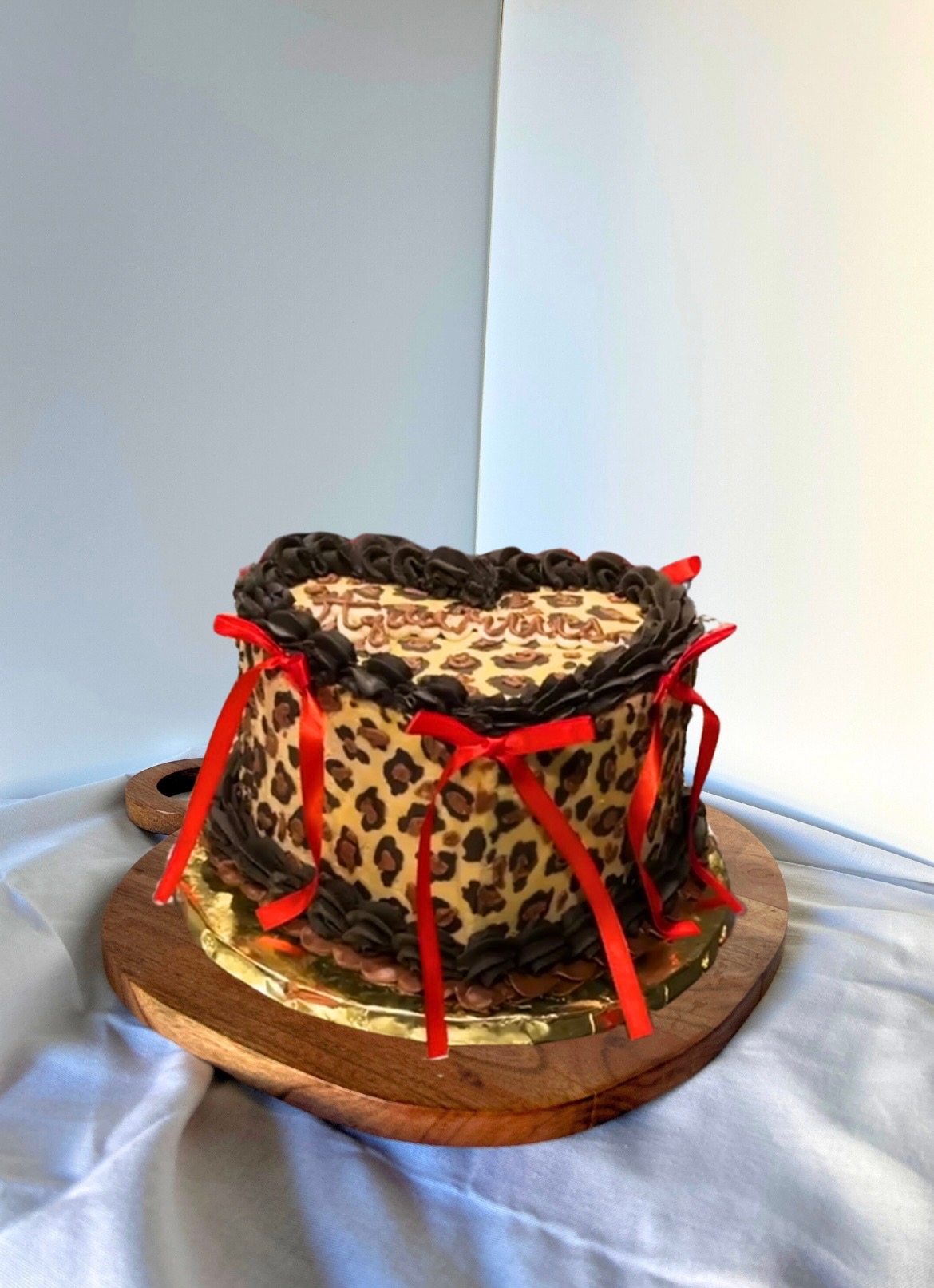 Heart-shaped leopard-print cake with chocolate piping and red ribbon bows on a wooden stand.