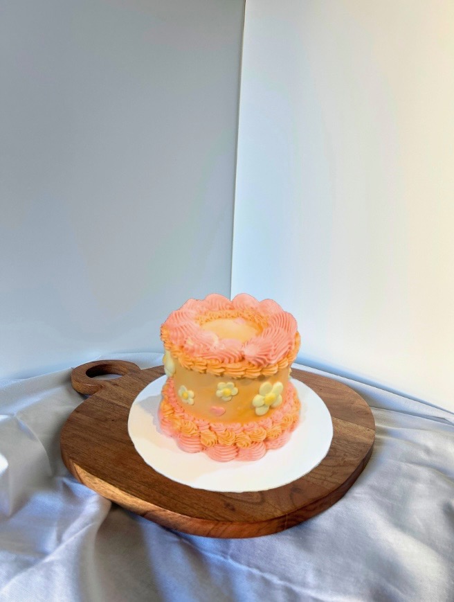 Peach buttercream cake with pink piped borders and small daisy accents on a wooden stand.